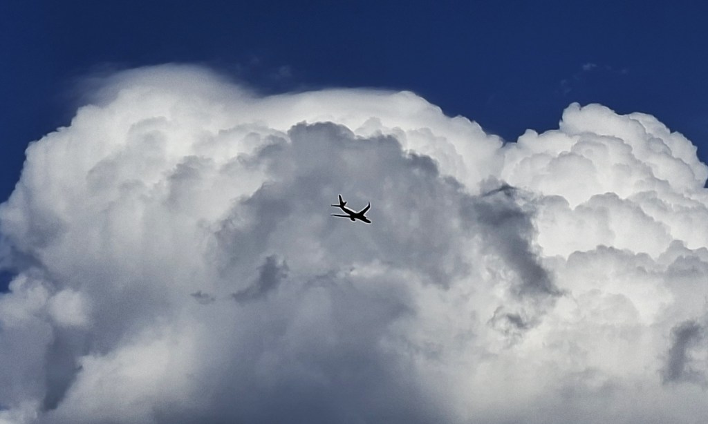 Blauer Himmel mit einer Wolke, in der Bildmitte in der Wolke ein Flugzeug